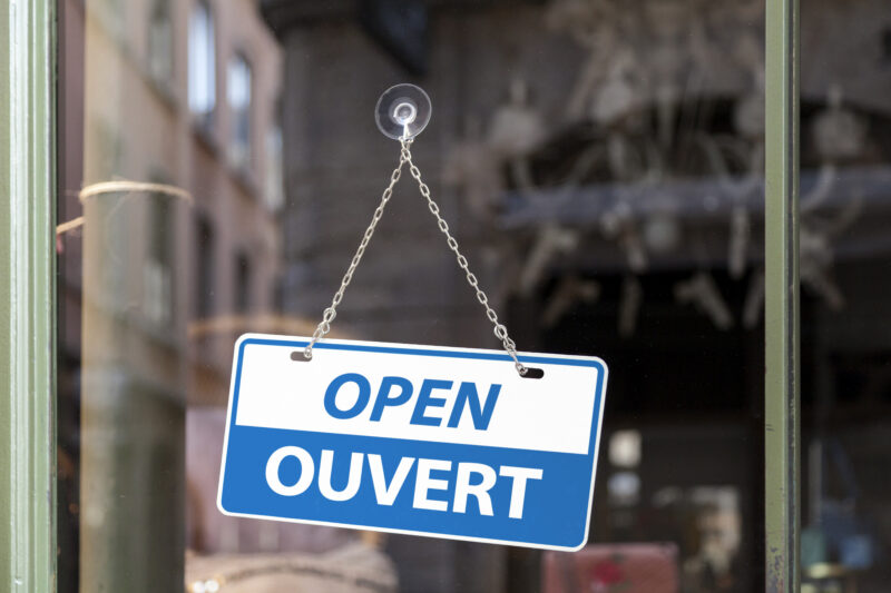 Close-up on a blue Open sign in a window with written in it in English & French.